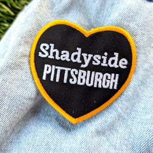May include: A heart-shaped patch with the words "Shadyside PITTSBURGH" in white lettering on a black background. The patch has a yellow border and is attached to a light blue denim jacket.