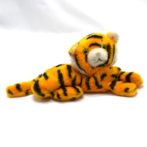May include: A plush toy tiger in a lying position. The tiger has orange fur with black stripes, a white face, and green eyes. The toy is made of soft material and is designed to resemble a tiger cub.