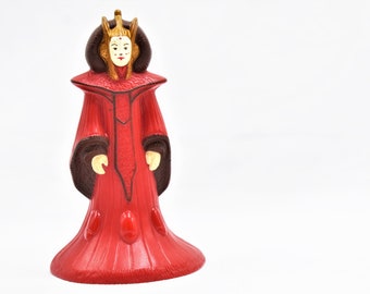 May include: A red plastic action figure of a queen wearing a gold crown and a red and brown robe.