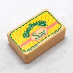 Può includere: Una saponetta vintage di Cabbage Patch Kids in un astuccio rettangolare. L'astuccio è beige con un'etichetta gialla e rosa con il logo Cabbage Patch Kids e la scritta "SOAP". Made in Italy.