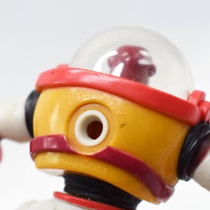 May include: Close-up of a yellow and red toy robot with a clear dome head. The robot has white arms and red accents. The text "&copy; Disney/Pixar" and "2002 China" are visible on the toy.