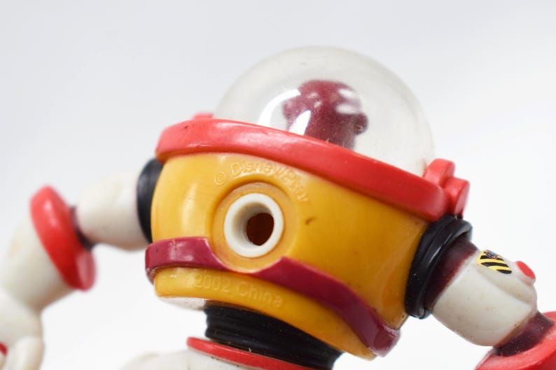 May include: Close-up of a yellow and red toy robot with a clear dome head. The robot has white arms and red accents. The text "&copy; Disney/Pixar" and "2002 China" are visible on the toy.