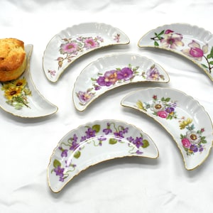 Puede incluir: Conjunto de platos de porcelana blancos en forma de media luna con diseños florales y ribetes dorados. Un plato contiene un bollo dorado. Los otros platos presentan varios patrones florales en rosa, morado y amarillo.
