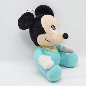 Op de afbeelding: Een pluche Mickey Mouse-speelgoed in een zittende positie. Het speelgoed heeft zwarte oren, een zwarte neus en een lichtbruin gezicht. Het draagt een lichtblauwe outfit met een gele kraag en witte handschoenen. Het speelgoed is ongeveer 30 cm hoog.
