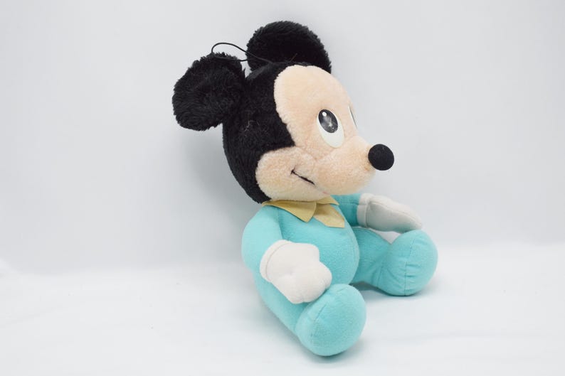 Op de afbeelding: Een pluche Mickey Mouse-speelgoed in een zittende positie. Het speelgoed heeft zwarte oren, een zwarte neus en een lichtbruin gezicht. Het draagt een lichtblauwe outfit met een gele kraag en witte handschoenen. Het speelgoed is ongeveer 30 cm hoog.