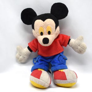 K&ouml;nnte beinhalten: Ein Vintage-Mickey-Mouse-Pl&uuml;schtier. Das Pl&uuml;schtier hat einen schwarzen Kopf mit gro&szlig;en Ohren, ein beiges Gesicht und ein rotes Oberteil. Es tr&auml;gt blaue Hosen, wei&szlig;e Handschuhe und rot-wei&szlig;-gelbe Schuhe. Das Spielzeug ist mit ausgestreckten Armen posiert.