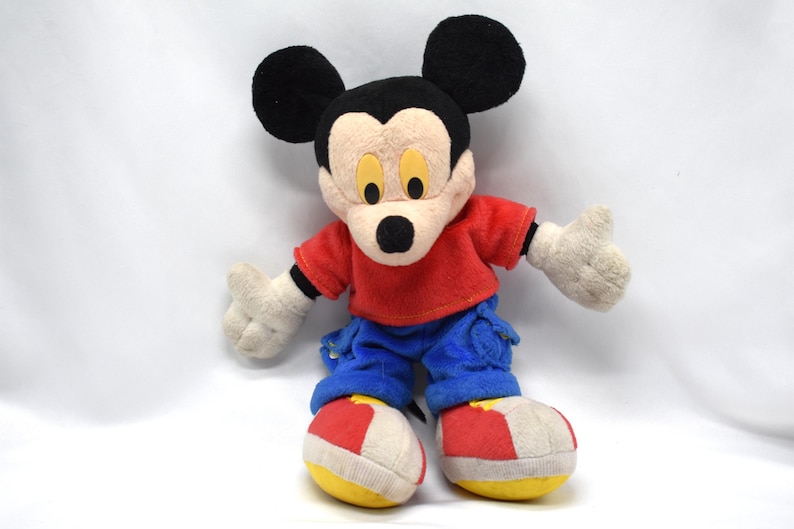 K&ouml;nnte beinhalten: Ein Vintage-Mickey-Mouse-Pl&uuml;schtier. Das Pl&uuml;schtier hat einen schwarzen Kopf mit gro&szlig;en Ohren, ein beiges Gesicht und ein rotes Oberteil. Es tr&auml;gt blaue Hosen, wei&szlig;e Handschuhe und rot-wei&szlig;-gelbe Schuhe. Das Spielzeug ist mit ausgestreckten Armen posiert.