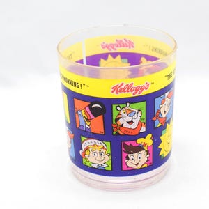 Op de afbeelding: Een doorzichtig plastic drinkglas met Kellogg's cartoonfiguren. Het glas heeft een gele band met het Kellogg's logo en de woorden "THE BEST MORNING!". Het glas is versierd met cartoonafbeeldingen in blauwe vierkanten.