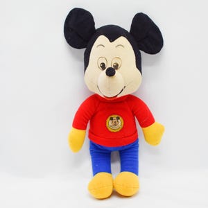May include: A plush Mickey Mouse doll. The doll has a black head with large ears, a tan face, and a black nose. It wears a red long-sleeved shirt, blue pants, and yellow gloves and shoes. A circular emblem is on the shirt.