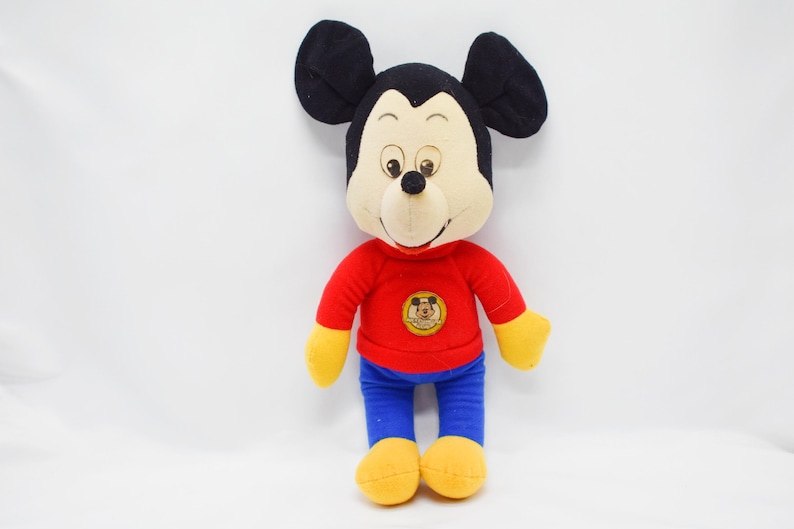 May include: A plush Mickey Mouse doll. The doll has a black head with large ears, a tan face, and a black nose. It wears a red long-sleeved shirt, blue pants, and yellow gloves and shoes. A circular emblem is on the shirt.