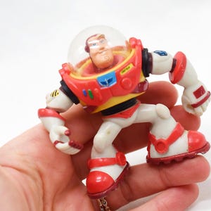 May include: A Buzz Lightyear action figure from the Toy Story franchise. The figure is primarily white, red, and yellow, with a clear plastic helmet. The toy is held in a hand, showcasing its poseable limbs and detailed design.