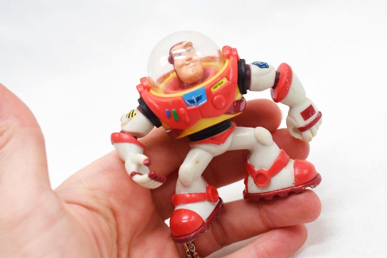 May include: A Buzz Lightyear action figure from the Toy Story franchise. The figure is primarily white, red, and yellow, with a clear plastic helmet. The toy is held in a hand, showcasing its poseable limbs and detailed design.