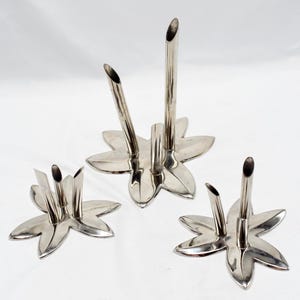 Op de afbeelding: Vier zilverkleurige bloemenvazen. Elke vaas heeft een stervormige basis met gebogen bloemblaadjes en een cilindrische buis voor het vasthouden van stelen. De vazen zijn gerangschikt op een wit oppervlak.