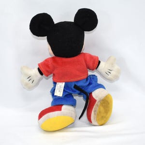 K&ouml;nnte beinhalten: Ein Mickey Mouse Pl&uuml;schtier, von hinten gesehen, mit schwarzen Ohren und Kopf, rotem Hemd, blauer Hose, wei&szlig;en Handschuhen und gelben, roten und wei&szlig;en Schuhen. Die Puppe steht vor einem wei&szlig;en Hintergrund.