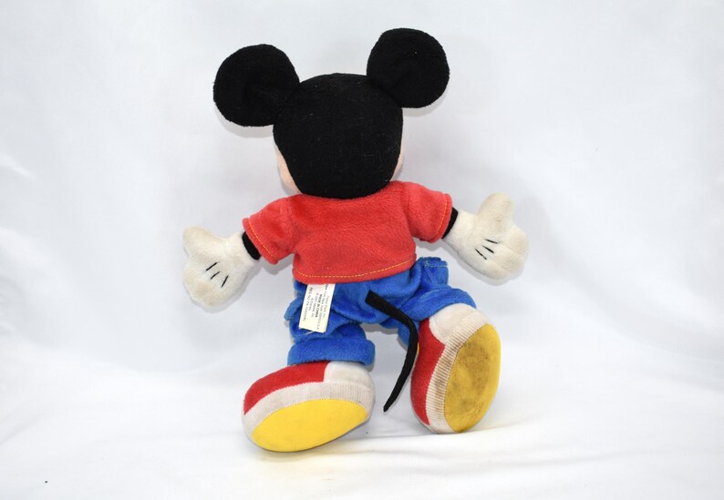 K&ouml;nnte beinhalten: Ein Mickey Mouse Pl&uuml;schtier, von hinten gesehen, mit schwarzen Ohren und Kopf, rotem Hemd, blauer Hose, wei&szlig;en Handschuhen und gelben, roten und wei&szlig;en Schuhen. Die Puppe steht vor einem wei&szlig;en Hintergrund.