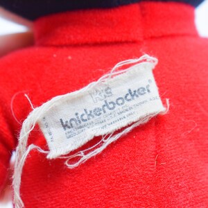May include: Close-up of a red plush toy with a black hat. A white tag with the text "knickerbocker" is attached to the toy. The tag also includes additional text. The toy is made of a soft, fuzzy material.