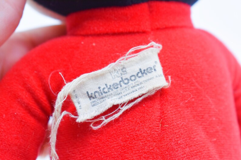 May include: Close-up of a red plush toy with a black hat. A white tag with the text "knickerbocker" is attached to the toy. The tag also includes additional text. The toy is made of a soft, fuzzy material.