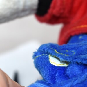 K&ouml;nnte beinhalten: Nahaufnahme eines Pl&uuml;schtiers in Blau, Rot und Wei&szlig;. Der blaue Teil hat gelbe N&auml;hte. Das Spielzeug hat eine weiche, flauschige Textur. Das Bild ist vor einem schlichten wei&szlig;en Hintergrund aufgenommen.