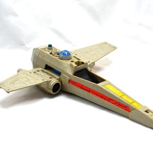 Peut inclure: Un jouet X-wing fighter beige et bleu avec des détails rouges et jaunes. Le jouet a des ailes détaillées et un cockpit. Un objet de collection Star Wars classique.