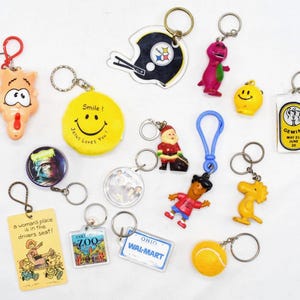 Może przedstawiać: Kolekcja breloków do kluczy o różnych wzorach. Zawiera żółtą uśmiechniętą buzię, hełm do futbolu amerykańskiego, dinozaura, figurkę Świętego Mikołaja i piłkę tenisową. Niektóre breloki mają tekst, w tym "Smile! Jesus Loves You!"