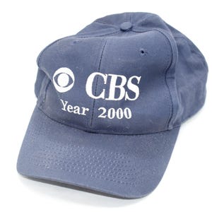 Könnte beinhalten: Marineblaue Baseballkappe mit dem CBS-Logo und dem Text "CBS Year 2000" in Weiß. Die Kappe hat einen gebogenen Schirm und einen Knopf oben. Der Hut ist aus weichem Stoff.
