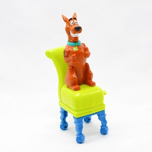 May include: A brown Scooby-Doo figurine sits on a lime green chair with blue legs. The cartoon dog is smiling and wearing a blue collar with a green tag. The chair has a curved back.