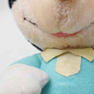 Op de afbeelding: Close-up van een vintage pluche pop met een lichtblauw shirt, witte handschoenen en een cr&egrave;mekleurige kraag. De pop heeft een lachend gezicht met zwarte ogen en een roze mond. De kop van de pop is zwart en cr&egrave;mekleurig.