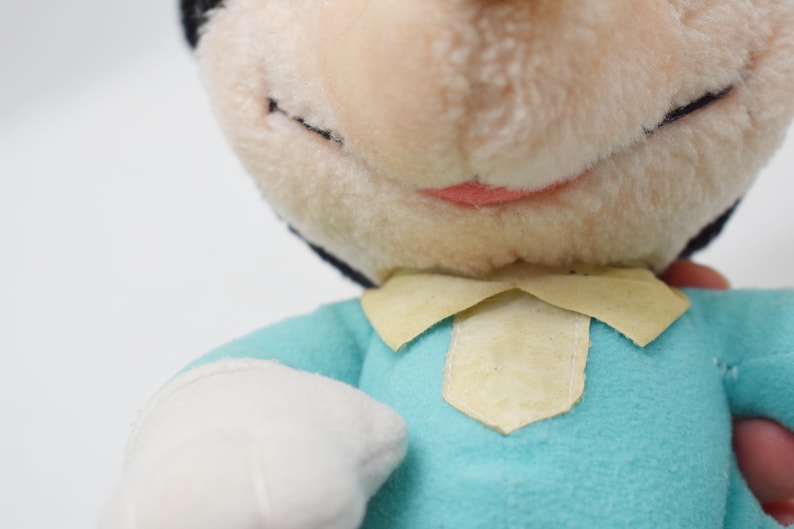 Op de afbeelding: Close-up van een vintage pluche pop met een lichtblauw shirt, witte handschoenen en een cr&egrave;mekleurige kraag. De pop heeft een lachend gezicht met zwarte ogen en een roze mond. De kop van de pop is zwart en cr&egrave;mekleurig.