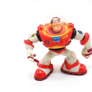 May include: A Buzz Lightyear action figure from the Toy Story franchise. The figure is primarily white with red and yellow accents. It features a clear helmet, a red chest plate, and red boots. The figure is in a crouching pose.