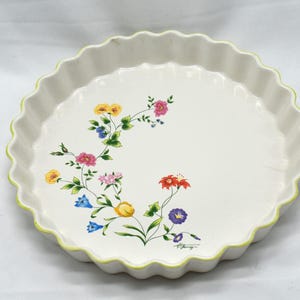 以下が含まれることがあります： 白いセラミック製のパイ皿。縁は波型で、花柄のデザイン。花は黄色、ピンク、赤、青、紫の色合い。縁には黄色のアクセント。