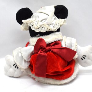 May include: A plush toy of a character with black ears, wearing a white bonnet with lace trim and a red dress with a large red bow. The character has white gloves with black details. The toy is on a white background.