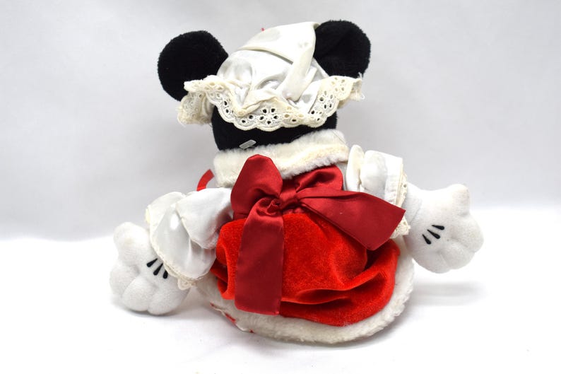 May include: A plush toy of a character with black ears, wearing a white bonnet with lace trim and a red dress with a large red bow. The character has white gloves with black details. The toy is on a white background.