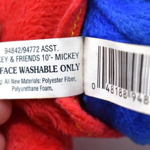 K&ouml;nnte beinhalten: Nahaufnahme eines Pl&uuml;schtiers mit roten und blauen Abschnitten. Ein wei&szlig;es Etikett lautet "MICKEY & FRIENDS 10"- MICKEY" und "SURFACE WASHABLE ONLY". Das Etikett listet auch F&uuml;llmaterialien auf. Ein Barcode ist sichtbar.