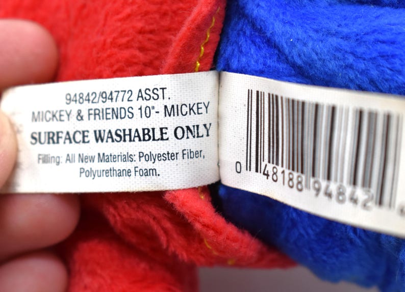 K&ouml;nnte beinhalten: Nahaufnahme eines Pl&uuml;schtiers mit roten und blauen Abschnitten. Ein wei&szlig;es Etikett lautet "MICKEY & FRIENDS 10"- MICKEY" und "SURFACE WASHABLE ONLY". Das Etikett listet auch F&uuml;llmaterialien auf. Ein Barcode ist sichtbar.