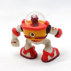 May include: A colorful toy robot with a clear dome head, yellow body, and red accents. The robot has white limbs and red shoes. The toy has a retro, space-age design, and is posed as if walking.