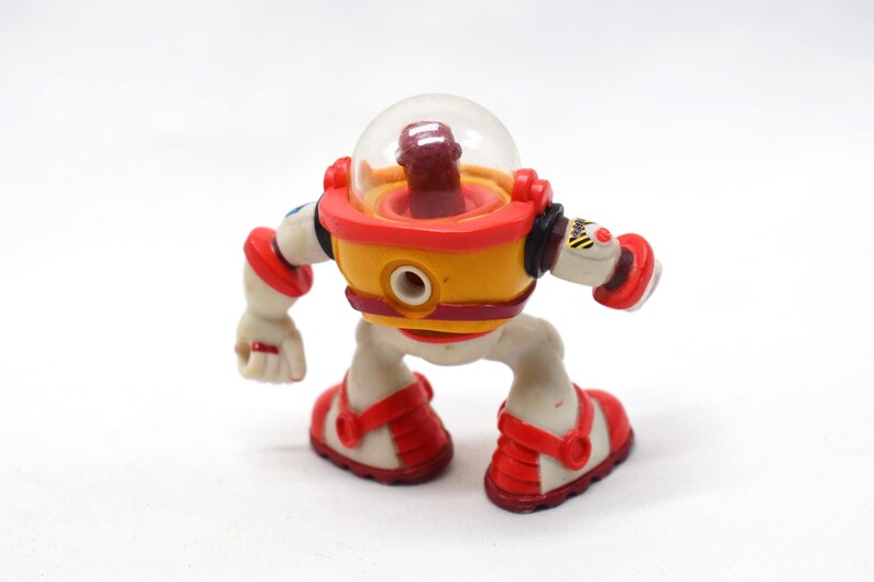 May include: A colorful toy robot with a clear dome head, yellow body, and red accents. The robot has white limbs and red shoes. The toy has a retro, space-age design, and is posed as if walking.