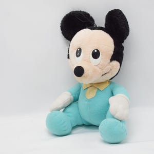 Op de afbeelding: Een pluche Mickey Mouse pop in een zittende positie. De pop heeft een lichtblauwe onesie, witte handschoenen en zwarte oren. Een gele vlinderdas zit om de nek. Het gezicht is beige met zwarte ogen en een zwarte neus.