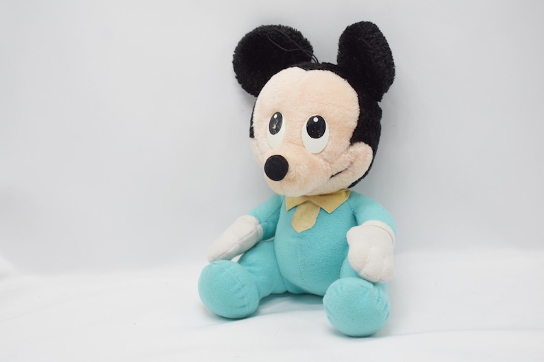 Op de afbeelding: Een pluche Mickey Mouse pop in een zittende positie. De pop heeft een lichtblauwe onesie, witte handschoenen en zwarte oren. Een gele vlinderdas zit om de nek. Het gezicht is beige met zwarte ogen en een zwarte neus.