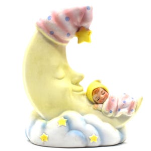 May include: A ceramic figurine of a sleeping baby nestled in a crescent moon. The moon is yellow with a smiling face, and the baby wears a yellow cap and pink pajamas with blue polka dots. A yellow star hangs from the moon.