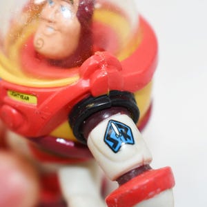 May include: A close-up of a Buzz Lightyear action figure. The figure is predominantly red, yellow, and white, with a clear helmet. The arm features a blue and white emblem. The word "LIGHTYEAR" is visible on the figure.
