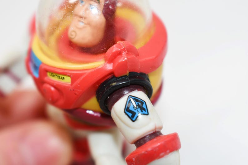 May include: A close-up of a Buzz Lightyear action figure. The figure is predominantly red, yellow, and white, with a clear helmet. The arm features a blue and white emblem. The word "LIGHTYEAR" is visible on the figure.
