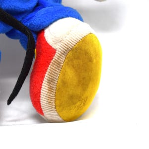 K&ouml;nnte beinhalten: Nahaufnahme eines Pl&uuml;schtierfu&szlig;es mit gelber Sohle, roter Seite und wei&szlig;em Rand. Das blaue Bein des Spielzeugs und ein schwarzer Riemen sind ebenfalls sichtbar. Das Spielzeug befindet sich auf wei&szlig;em Hintergrund.
