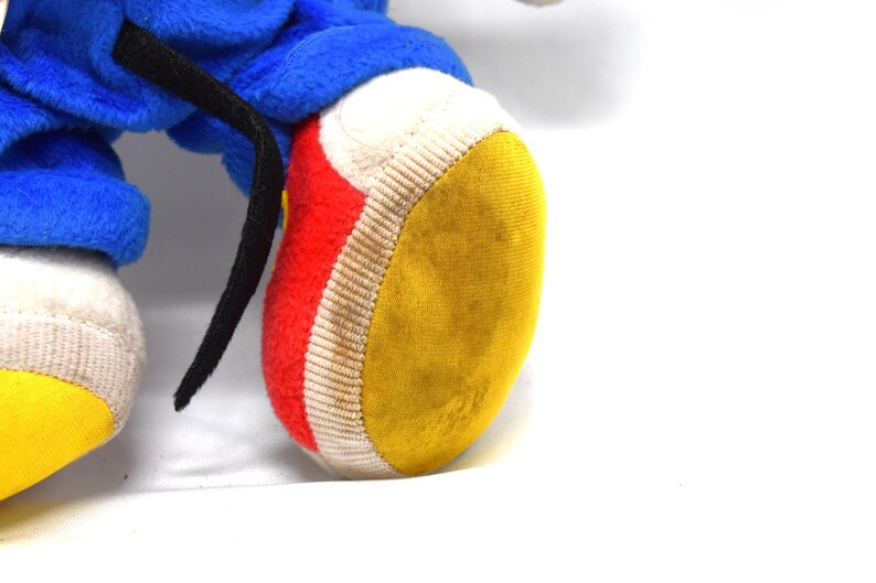 K&ouml;nnte beinhalten: Nahaufnahme eines Pl&uuml;schtierfu&szlig;es mit gelber Sohle, roter Seite und wei&szlig;em Rand. Das blaue Bein des Spielzeugs und ein schwarzer Riemen sind ebenfalls sichtbar. Das Spielzeug befindet sich auf wei&szlig;em Hintergrund.