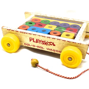 May include: A vintage Playskool Col-O-Rol wooden pull wagon featuring bright yellow plastic wheels and a red and white striped pull cord with a wooden bead. The wagon contains a set of colorful wooden blocks in red, green, blue, and yellow.