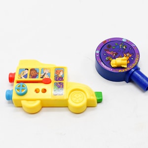 Op de afbeelding: Een gele speelgoed schoolbus met rode, blauwe en groene accenten, met stripfiguren in de ramen. Daarnaast een paars en blauw speelgoed met de tekst "Scholastic" en een geel object.