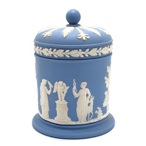 May include: A blue ceramic jar with a lid, featuring white bas-relief figures and floral designs. The jar has a round base and a knob on top. The figures depict classical scenes.
