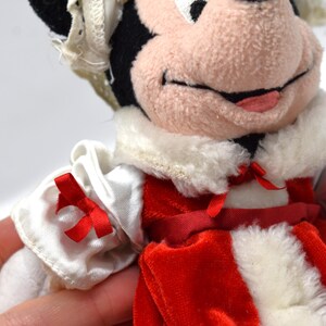 May include: A plush Minnie Mouse doll wearing a red and white dress with a white collar and red bows. The doll has black ears, a pink nose, and a smiling expression. The doll is a collectible item.