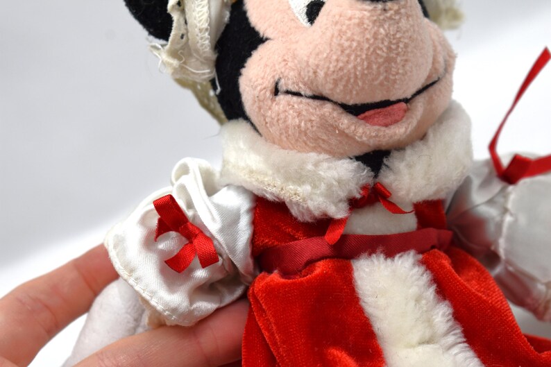 May include: A plush Minnie Mouse doll wearing a red and white dress with a white collar and red bows. The doll has black ears, a pink nose, and a smiling expression. The doll is a collectible item.