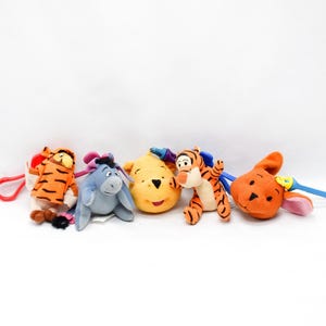 May include: A collection of plush toys featuring characters from Winnie the Pooh. The toys include Tigger, Eeyore, Winnie the Pooh, and Roo. Each toy is brightly colored and has a loop for hanging.
