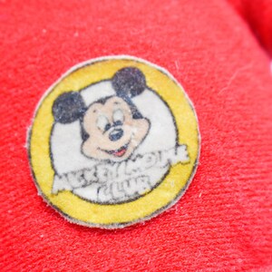 May include: Close-up of a red fabric with a circular Mickey Mouse Club patch. The patch features a smiling Mickey Mouse in a yellow circle with the words "Mickey Mouse Club".